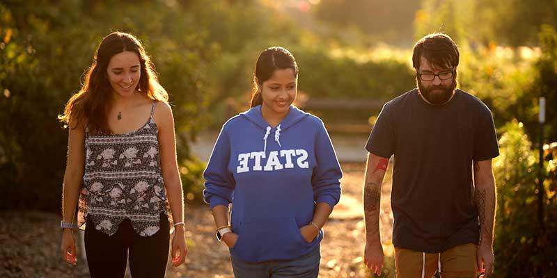 Three individuals outside with trees in the background. On the left is a man with brown hair and a matching beard and mustache. He wears a grey T-shirt. In the middle is a woman with brown hair pulled back in a ponytail. She wears a blue STATE hoodie. On the right is a woman with brown hair. She wears a black-and-white patterned tank top.