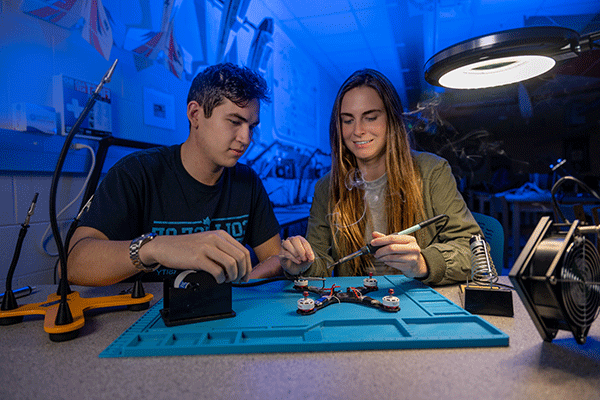 Two students are working in a blue-lit laboratory space. On the left is a white female student with long straight brown hair. She wears a dark green jacket with a grey undershirt. On the right is a white male student with short brown hair. He wears a black T-shirt with blue lettering on the front. The woman holds a soldering iron. Technology equipment is visible on the table in front of them with a blue tray under the equipment. A lamp light sits left of the equipment.