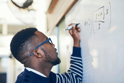 A Black man wearing glasses and a blue-and-white striped sweater writes a mathematical equation on a white board in a classroom or office setting.